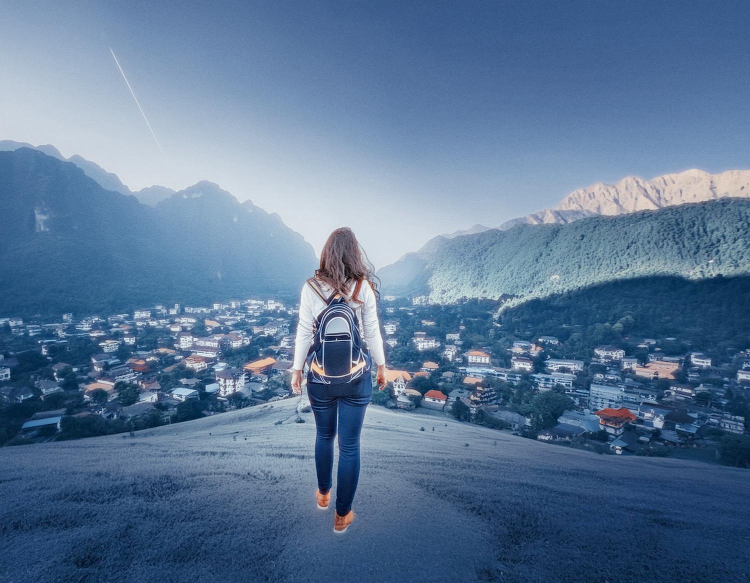 A stylish traveler with flowing hair in a chic jacket, jeans, and boots stands on a hill. Herschel Roll Top Backpack over one shoulder, they admire a town in the valley below and the clear sky above.