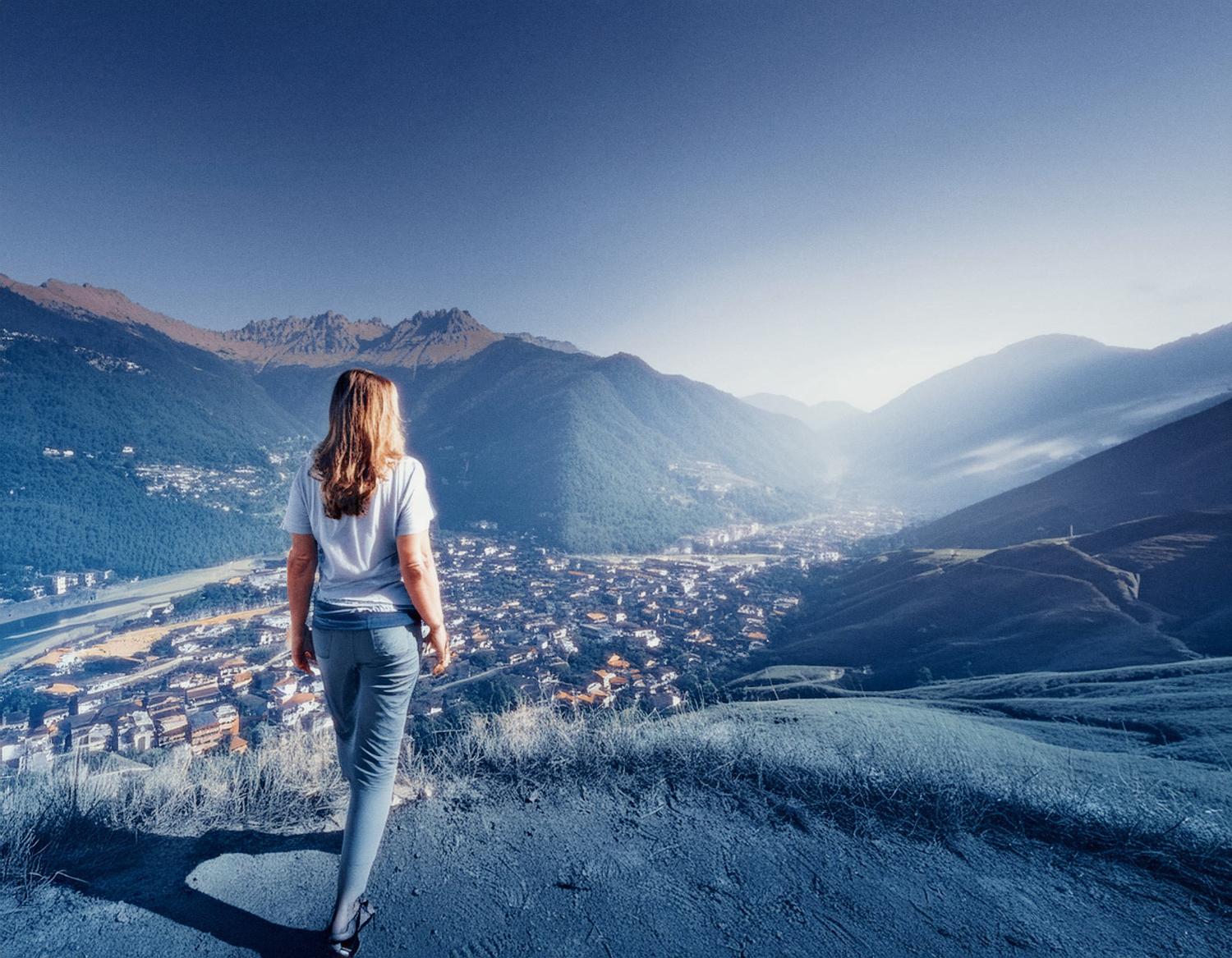 A woman with flowing hair stands on a hillside, overlooking a serene valley town basked in soft sunlight. Dressed casually in a white top and jeans, she carries a stylish Timbuk2 Spark Mini Backpack, perfect for her fashion-forward travels.