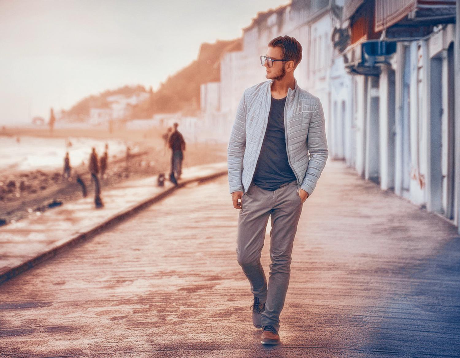 A man in chic sunglasses, a light jacket, and gray pants strolls a beachfront promenade. His sleek Samsonite brief backpack completes the effortlessly stylish look.