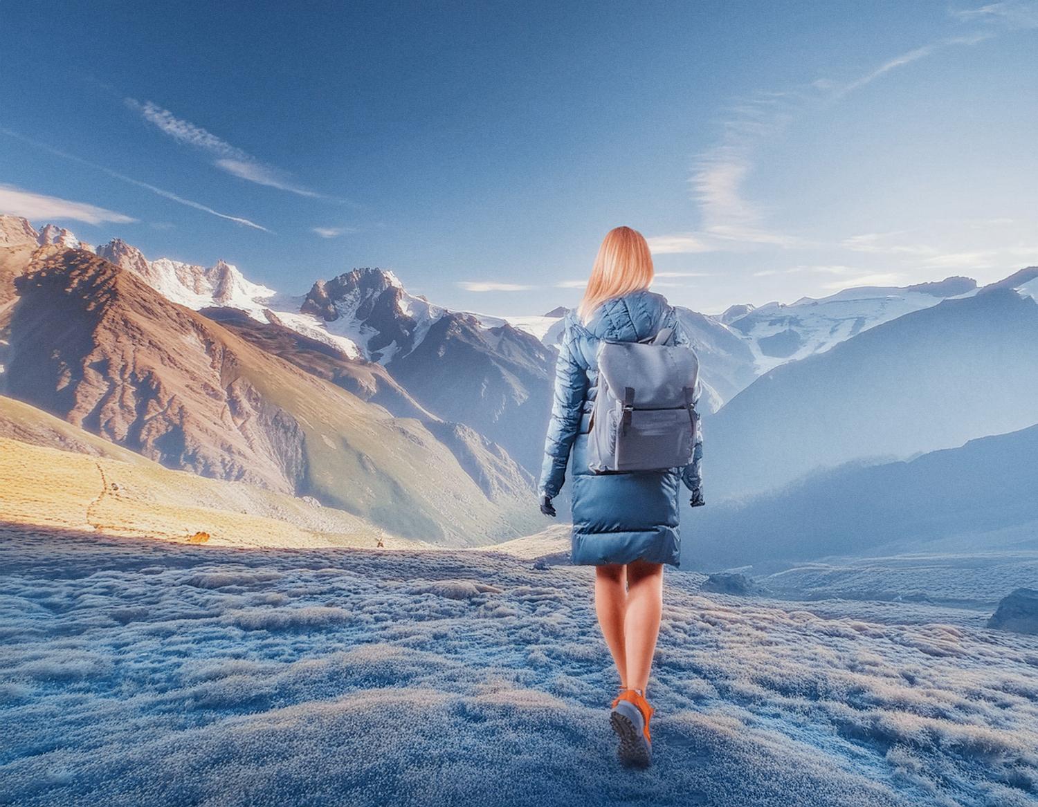 A fashion-forward traveler in a stylish blue coat pairs a sleek gray Samsonite backpack, striding across frost-kissed terrain towards majestic, snow-capped mountains under a pristine sky.
