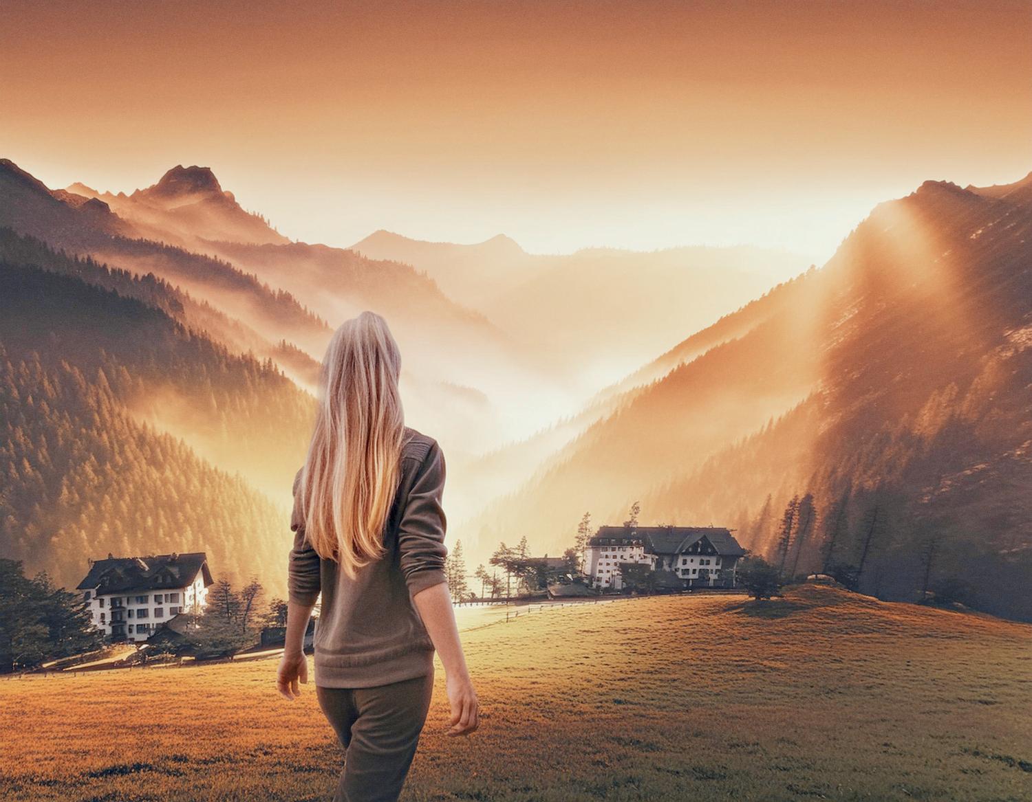 A person with long blonde hair stands in a grassy field, facing a picturesque sunrise over mountains. Sunlight filters through the mist, illuminating the valley and small houses below. Sporting the chic Timbuk2 Vapor Sling Crossbody Bag, they are all set for stylish adventures.