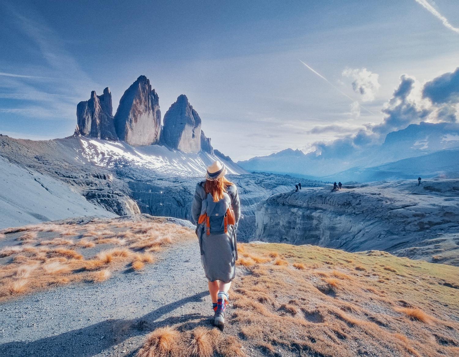 A stylish adventurer in a hat, grey dress, and Osprey Packs backpack strides through rocky terrain towards snow-capped peaks under a vibrant sky. Others explore the breathtaking mountainous landscape in the background. #TravelStyle