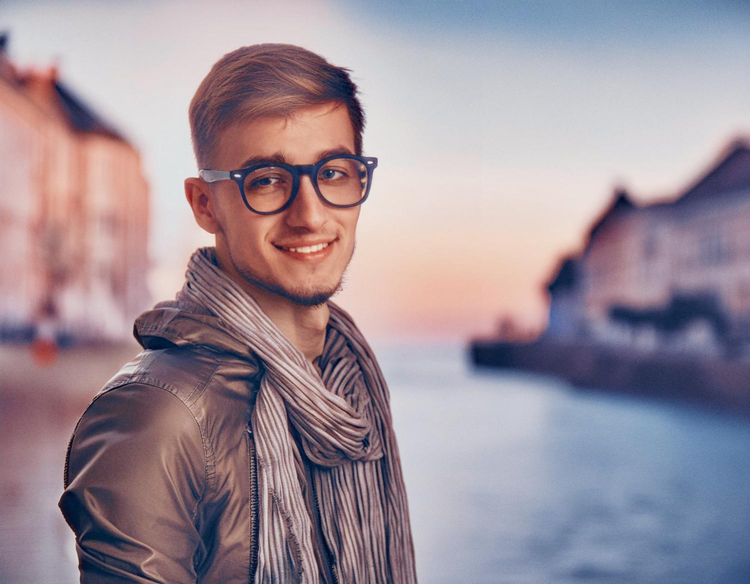 A stylish young man with chic blue glasses, a light brown jacket, and a gray scarf poses with an Osprey Packs Heritage Waist Pack against a sunset waterfront backdrop.