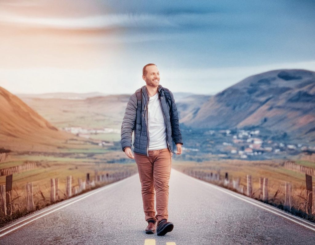 A bearded man in chic outdoor wear, sporting an Osprey Pack, strides down a serene road lined with fences towards a rustic valley.
