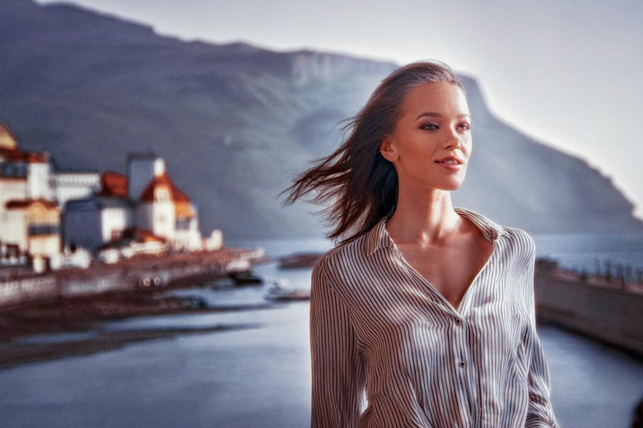 A woman in a classic striped button-up gazes over a serene coastal town, her hair tousled by the breeze. Timeless style meets tranquil beauty.