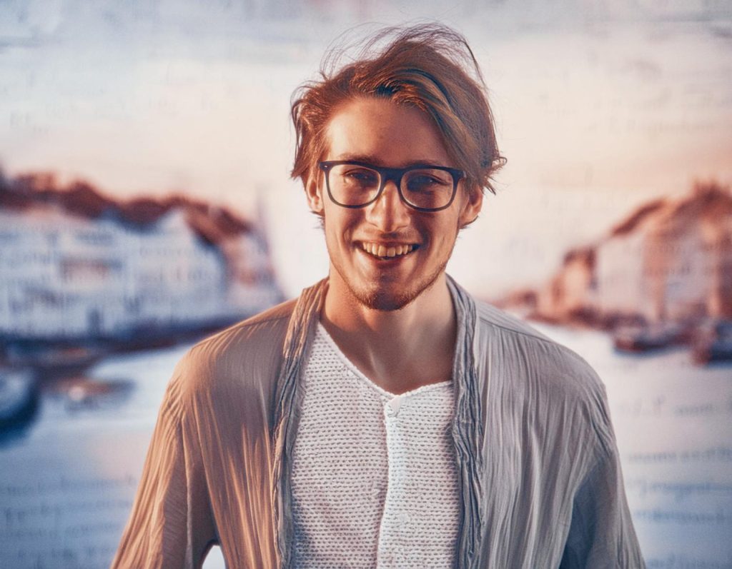 A stylish young man with glasses and tousled hair smiles at the camera, donning a textured shirt and relaxed cardigan. His JanSport Pro Crossbody Bag adds a touch of casual, modern flair against a softly blurred waterfront town backdrop.