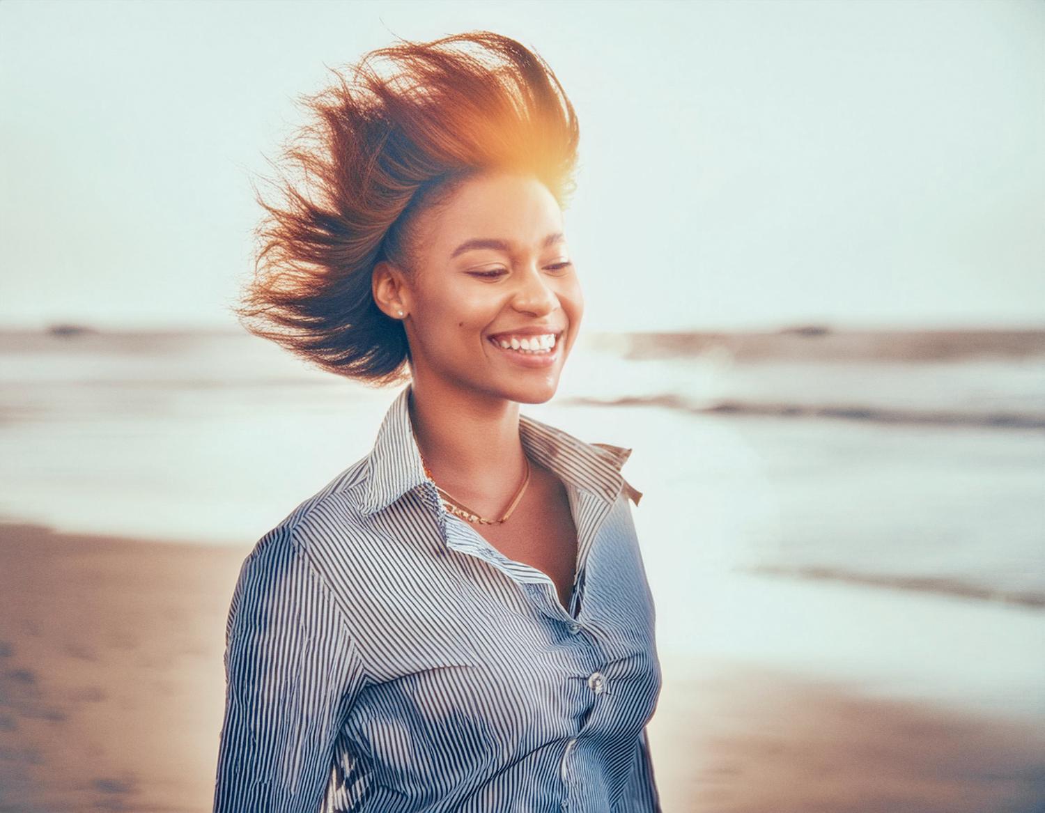 A joyful woman, her short hair windswept, stands on a beach at sunset. She radiates style in a light blue striped button-down and JanSport Pro Crossbody Bag, perfectly blending fashion and function.