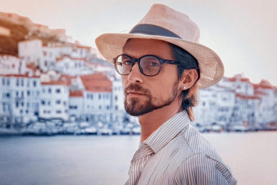 A bearded man with stylish glasses and a wide-brimmed hat poses before a scenic coastal town, showcasing his Heritage Watches bracelet. Trendy elegance!