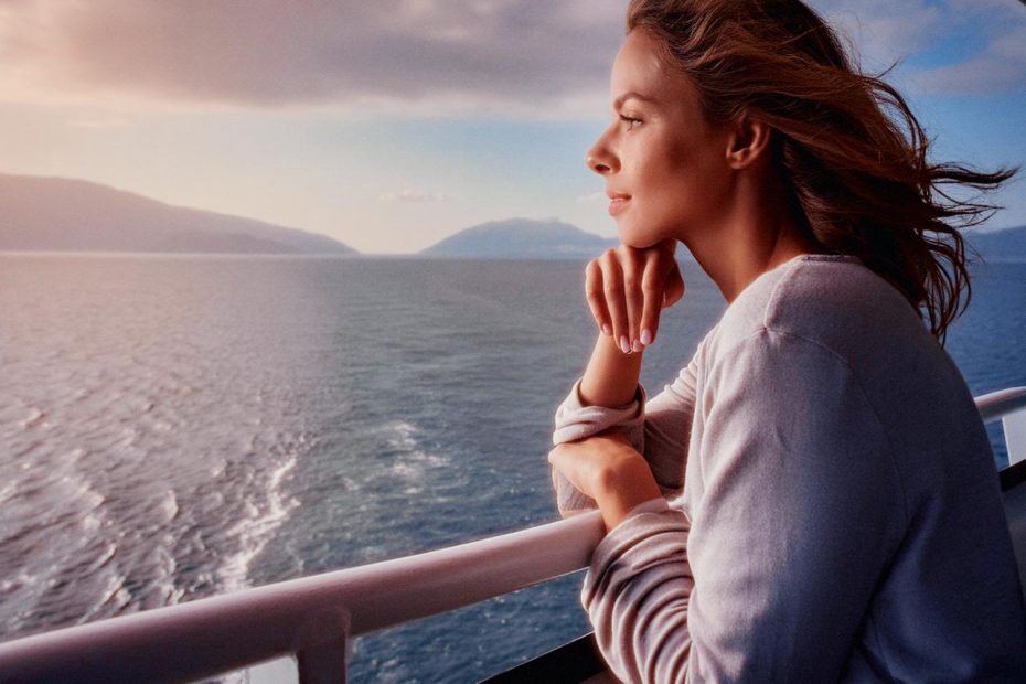 A woman in a light sweater leans on a boat railing, her Tumi Bravo bag by her side. She gazes at the calm sea and distant mountains, dreaming of stylish future travels.