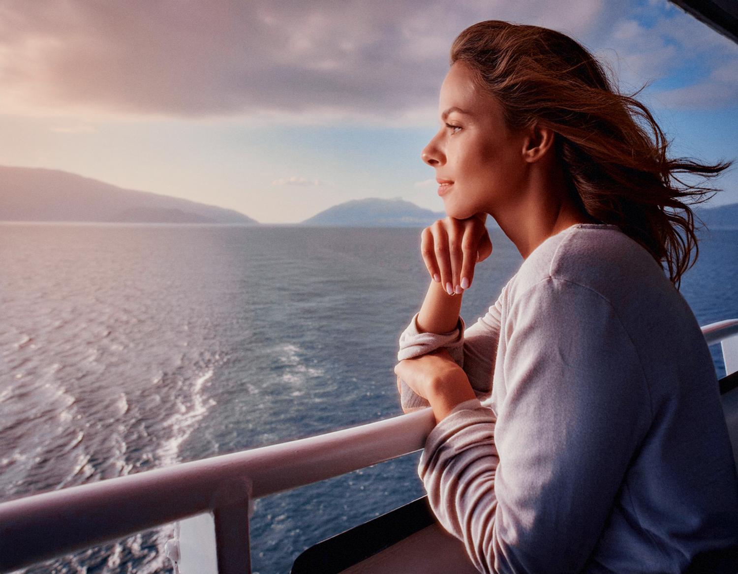 A woman in a light sweater leans on a boat railing, her Tumi Bravo bag by her side. She gazes at the calm sea and distant mountains, dreaming of stylish future travels.