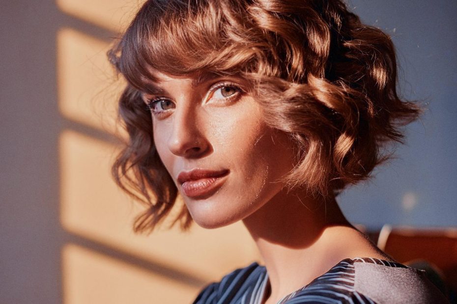 A woman with chic, wavy brown hair and bangs gazes confidently at the camera. Her dark striped top exudes timeless Parisian style, paired perfectly with moody lighting that casts dramatic shadows behind her.