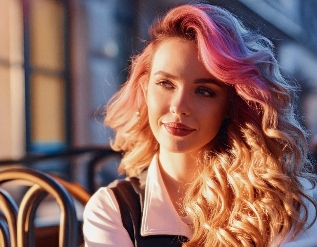 A woman with long, wavy blonde hair dipped in pink highlights smiles softly. In a cozy indoor setting, she sports a chic white top beside her stylish London Roll-top Backpack. Warm light and blurred wooden chairs create an inviting backdrop.