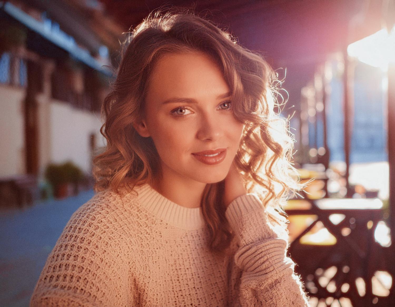 A chic woman with long, wavy hair sits outdoors in soft sunlight, wearing a light sweater. A Herschel Supply classic bag by her side, she smiles calmly amid blurred tables and buildings.