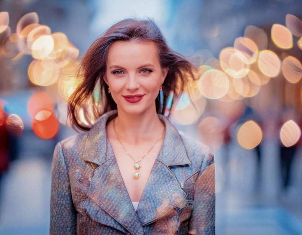 A woman with long brown hair smiles subtly in an urban evening scene, donning a chic patterned coat, pendant necklace, and Herschel Supply backpack. Bokeh lights twinkle behind her.