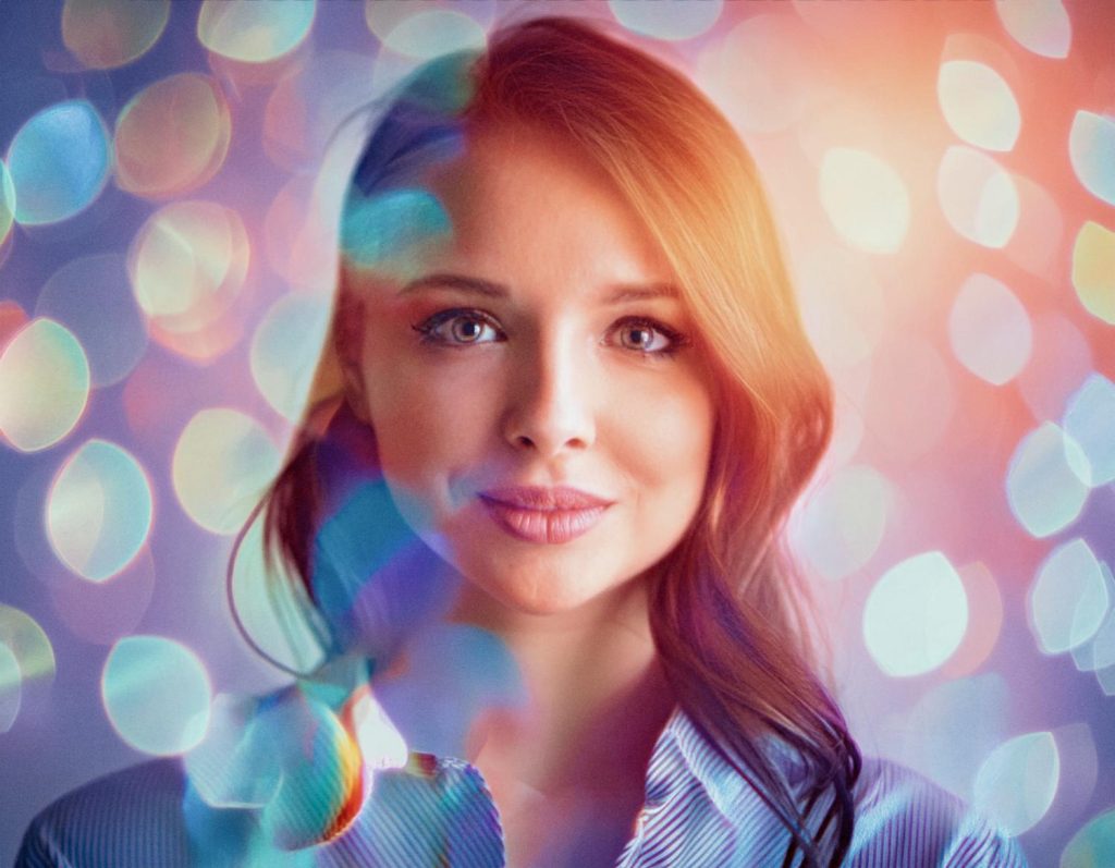 A woman with long, wavy auburn hair and a light blue button-up shirt holds a chic suede JanSport backpack against vibrant bokeh lights. Trendy and captivating.