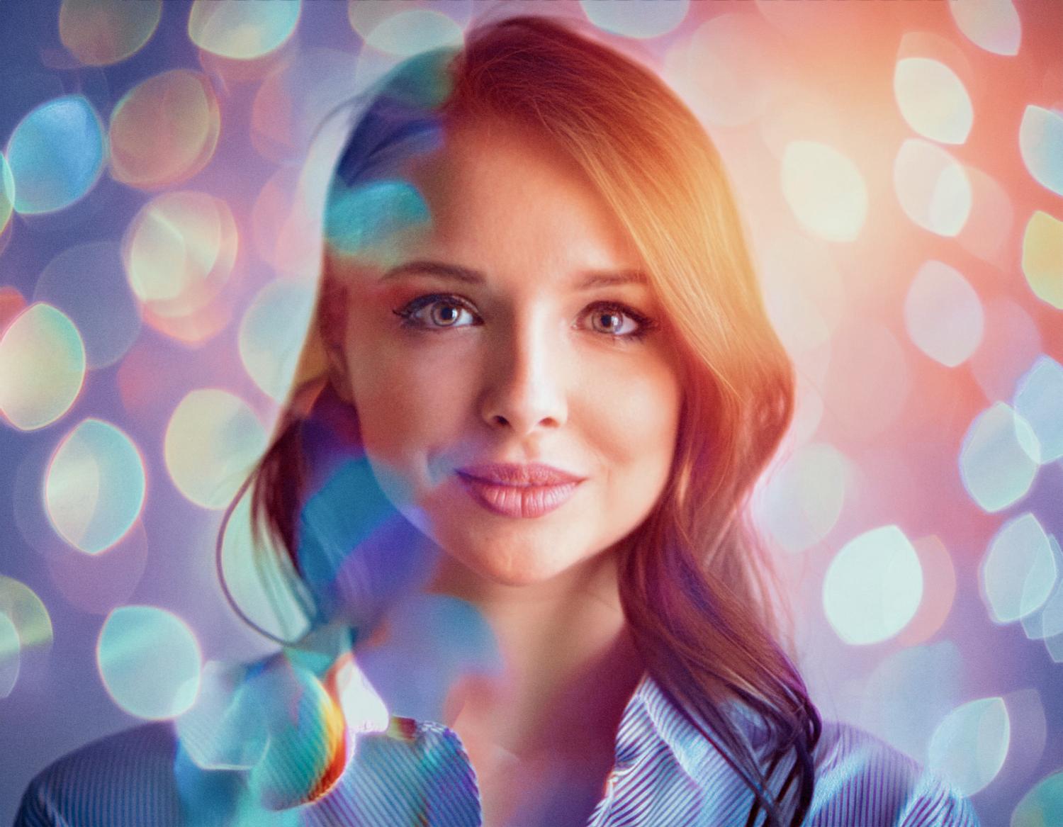 A woman with long, wavy auburn hair and a light blue button-up shirt holds a chic suede JanSport backpack against vibrant bokeh lights. Trendy and captivating.