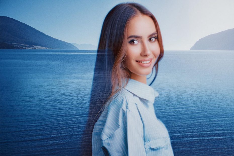 A stylish young woman with long brown hair and a light blue shirt smiles, showcasing her chic INCASE Transfer Two-Way Tote against a serene backdrop of calm water and mountains.
