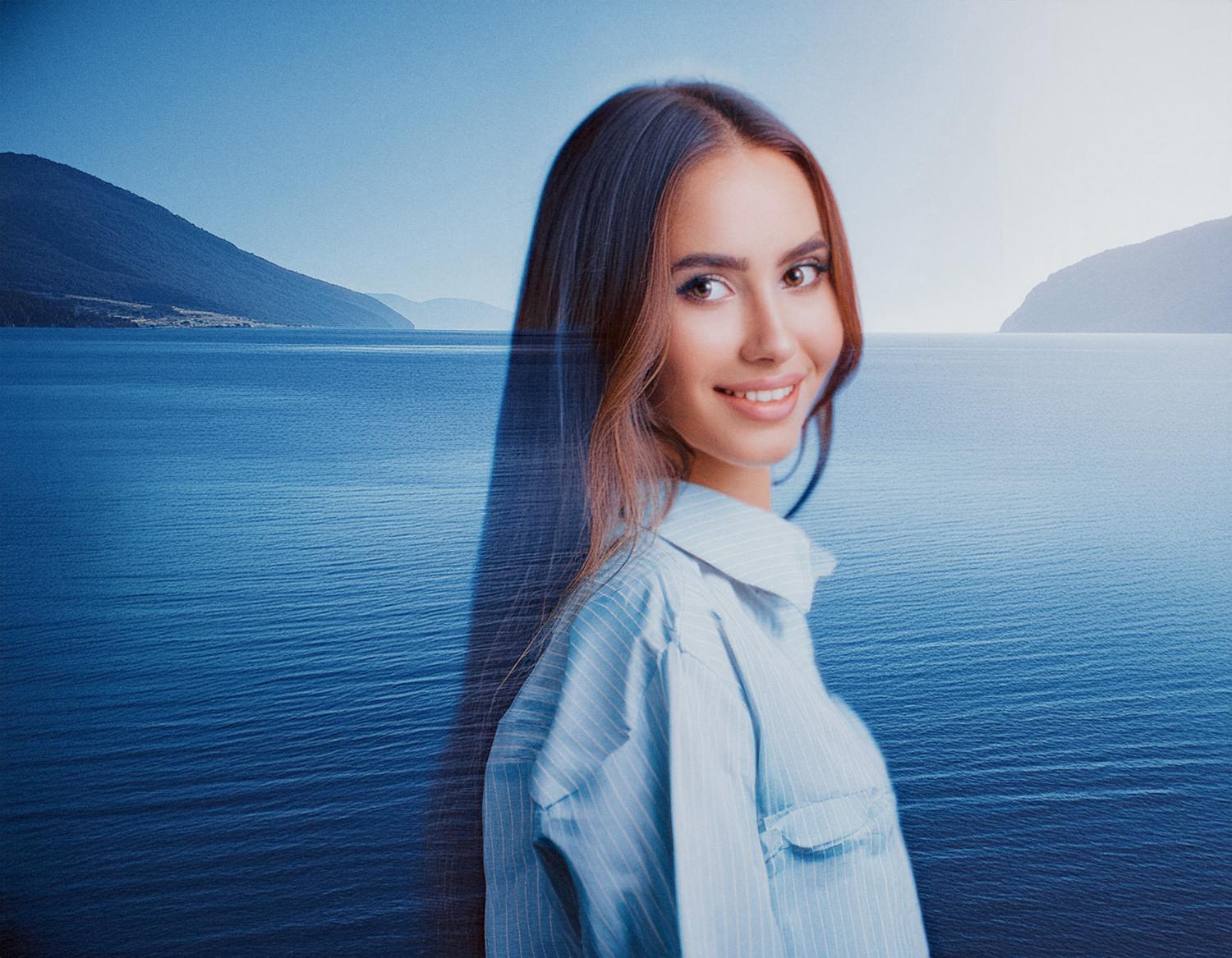 A stylish young woman with long brown hair and a light blue shirt smiles, showcasing her chic INCASE Transfer Two-Way Tote against a serene backdrop of calm water and mountains.