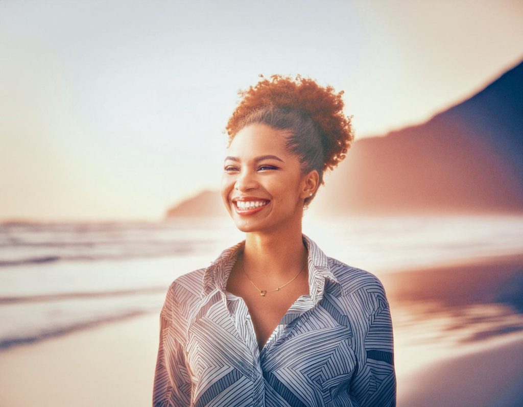 A woman with curly hair, radiant smile, and patterned shirt stands on a beach at sunset, sporting a JanSport backpack. Soft waves and mountains in the background. #BeachFashion