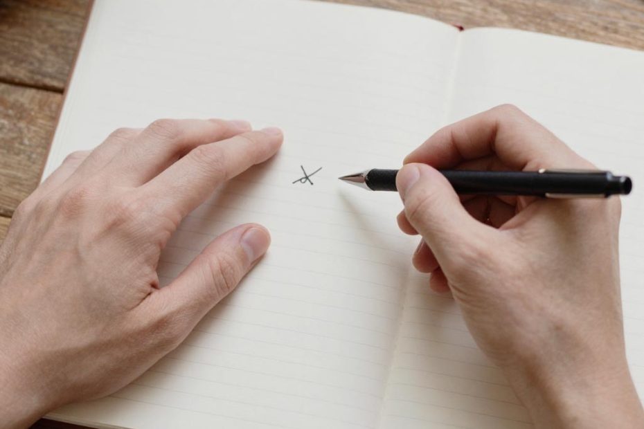 A person sketches a small X in a lined notebook on a wooden table, brainstorming trendy DIY bag charm ideas for stylish accessories.